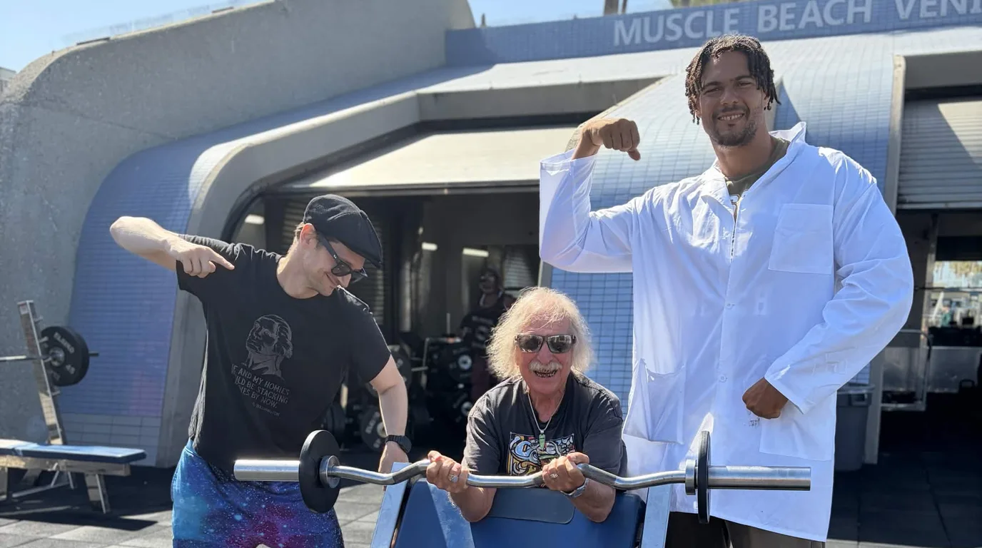Three people at the Muscle Beach Venice outdoor gym under the MUSCLE BEACH VENICE sign — one standing at left in galaxy-print trousers and a black science T-shirt, pointing down at a curl bar; one seated mid-curl at a preacher bench; one in a white lab coat flexing at right